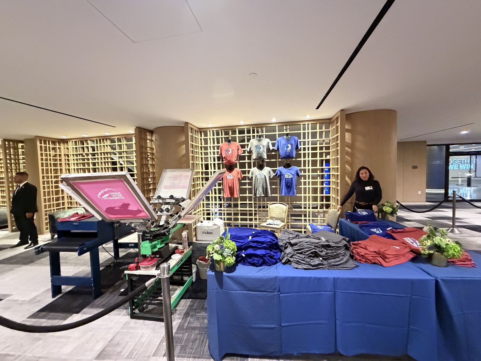 Expansive trade show booth with illuminated gridwall display, printed apparel on table, and multiple visitors browsing merchandise