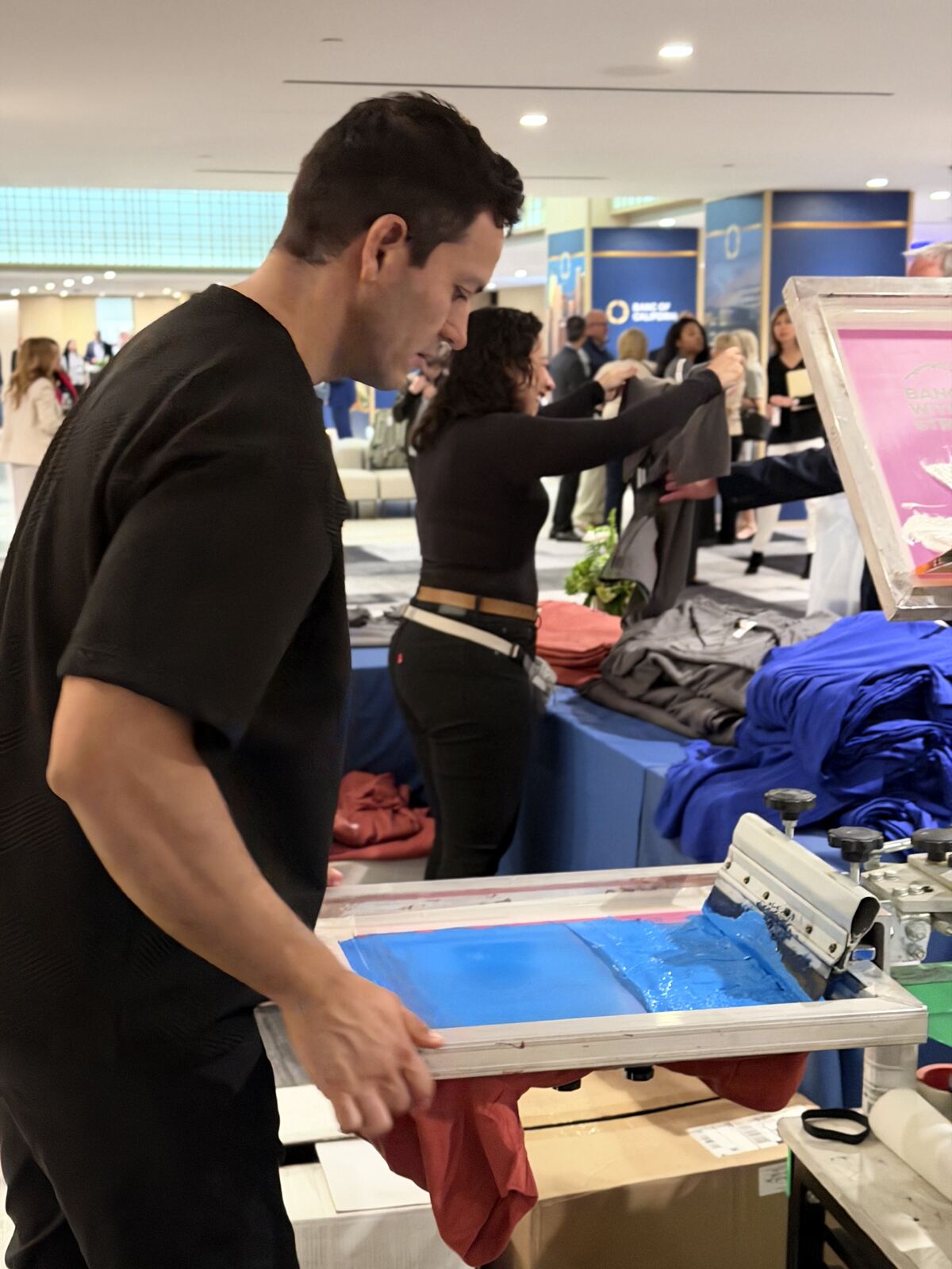 Person using screen-printing equipment at booth table with audience in background